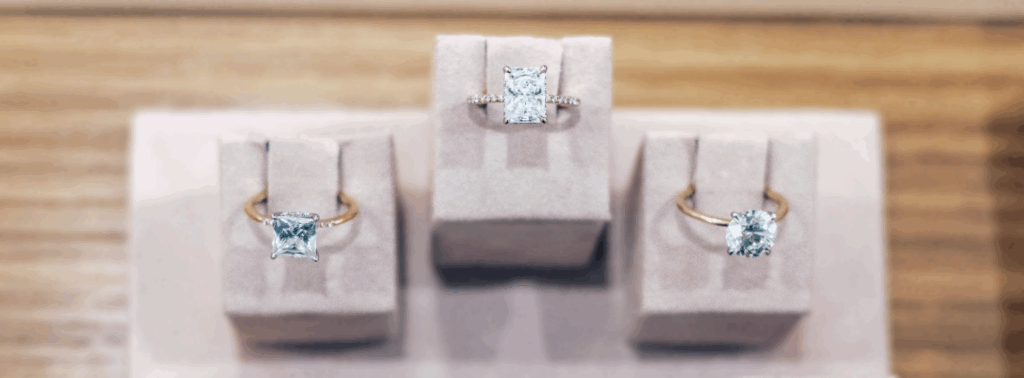 Three diamond rings are displayed on beige velvet stands. The rings have gold bands and prominent diamond settings, with the center ring featuring additional small diamonds along its band. The background is wood-toned.