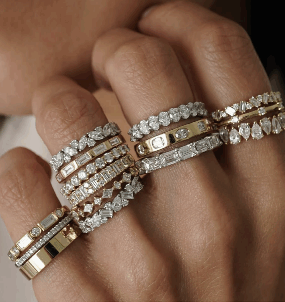 A close-up of a hand with four fingers covered in stacked gold, silver, and diamond rings, showcasing various sparkling designs and cuts.