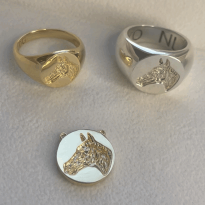 The Equestrian Line features gold and silver signet rings with a matching round pendant, each engraved with a detailed horse head, displayed on a light surface.