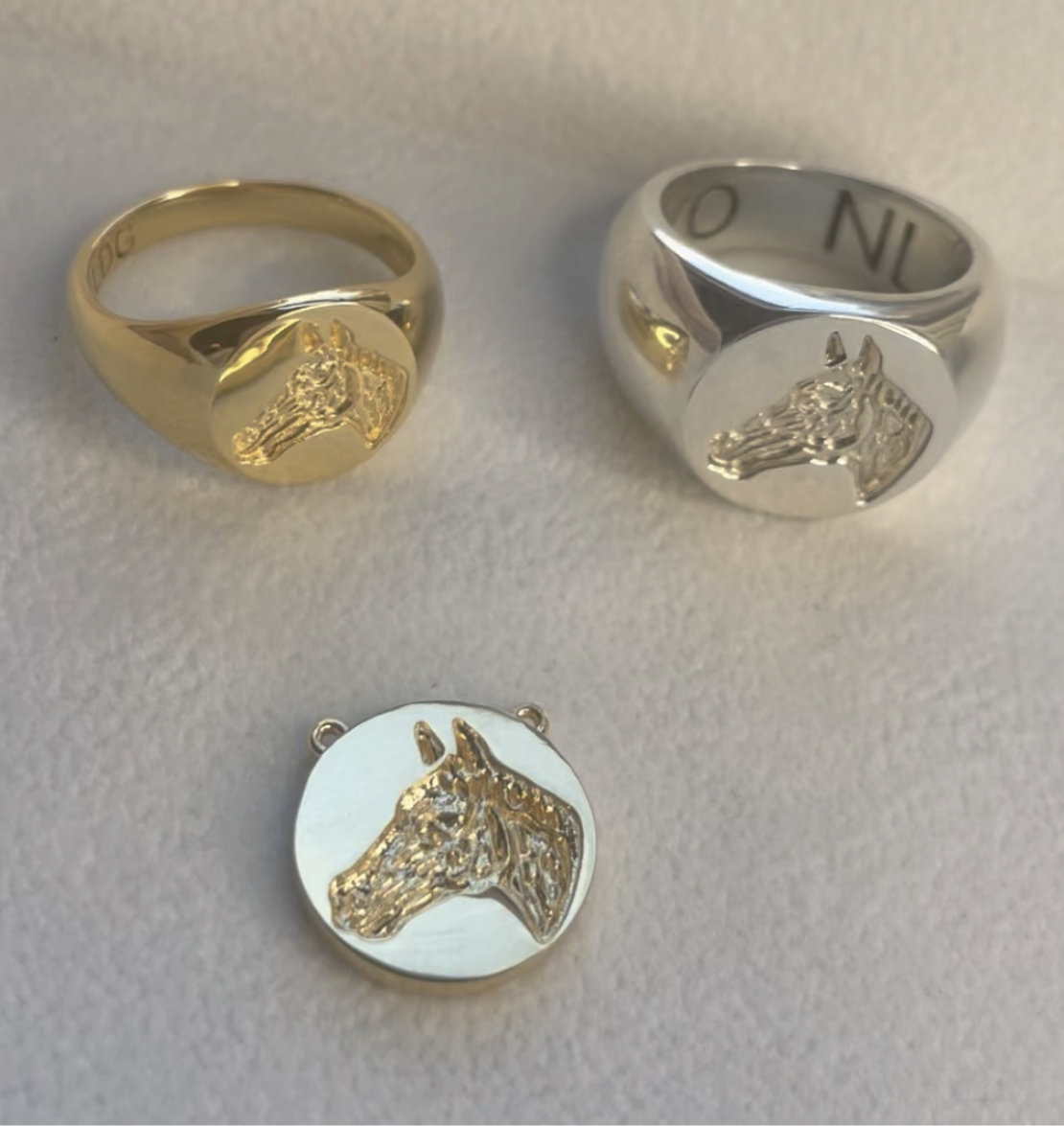 The Equestrian Line features gold and silver signet rings with a matching round pendant, each engraved with a detailed horse head, displayed on a light surface.