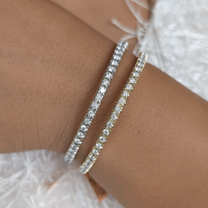 product Two Tennis Bracelets, one in white metal and one in yellow metal, each adorned with round-cut diamonds, are shown on a wrist against a textured white background.