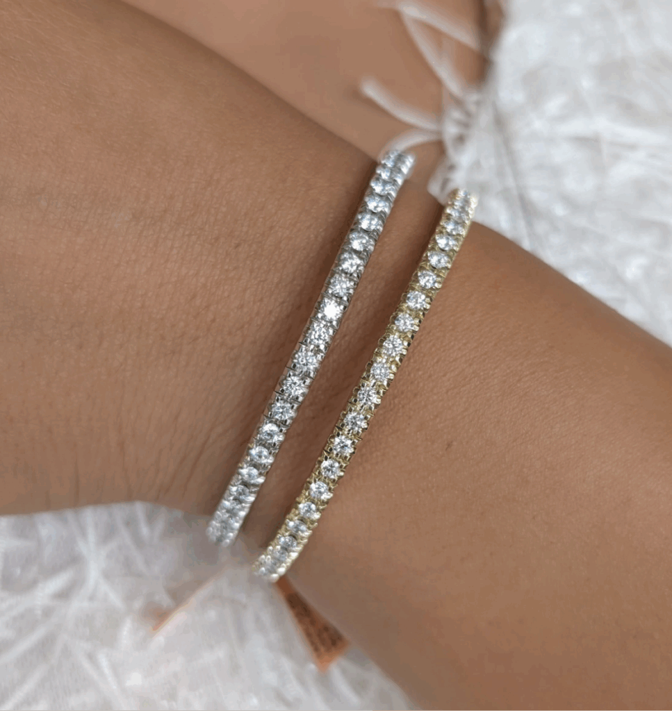 product Two Tennis Bracelets, one in white metal and one in yellow metal, each adorned with round-cut diamonds, are shown on a wrist against a textured white background.